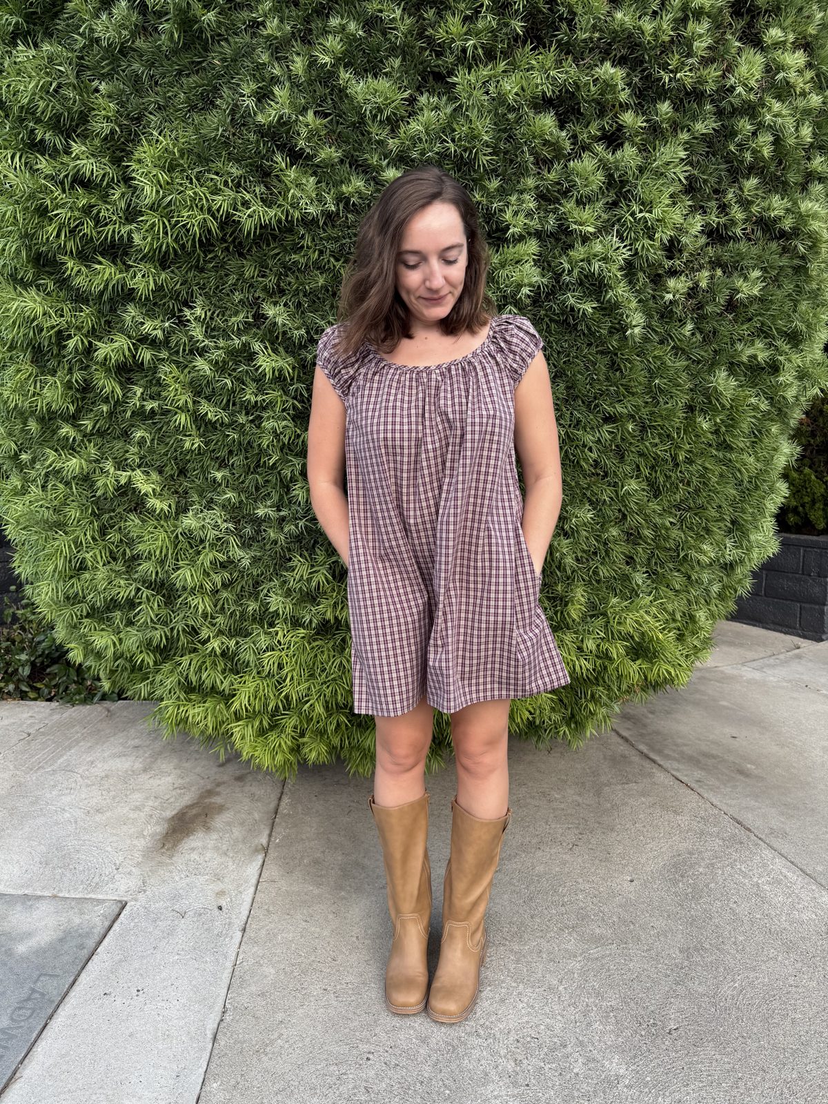 Emily smiling in her plaid Linnea mini dress in front of a hedge